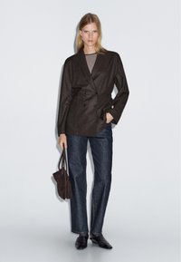 Žena v tmavém dvouřadém blejzru, pruhovaném topu, tmavých rovně střižených džínách, černých botách a držící hnědou kabelku na jednobarevném pozadí.