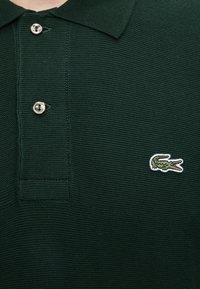 Polo vert foncé texturé avec deux boutons blancs et un petit logo crocodile vert brodé sur la poitrine gauche.