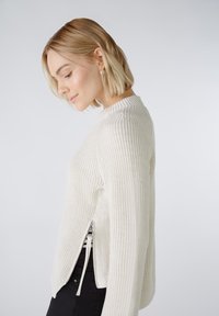 Texturierter cremefarbener Strickpullover mit Rundhalsausschnitt, seitlichen Reißverschluss-Schlüssen und gerippten Details, getragen über schwarzen Hosen.