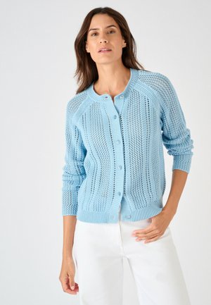 Mujer con un cárdigan de punto azul claro abotonado con cuello redondo y mangas remangadas, combinado con pantalones blancos de talle alto.