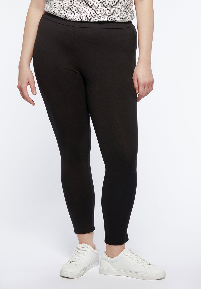 Leggings neri in tessuto elasticizzato, aderenti alla caviglia, abbinati a sneaker bianche, indossati con una maglietta stampata.