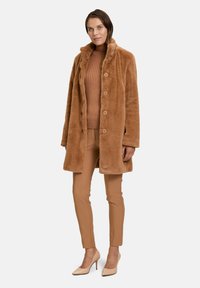 Faux fur brun frakke med krave og knaplukning, parret med en ribbet brun sweater og beige slim-fit bukser, afsluttet med beige hæle.