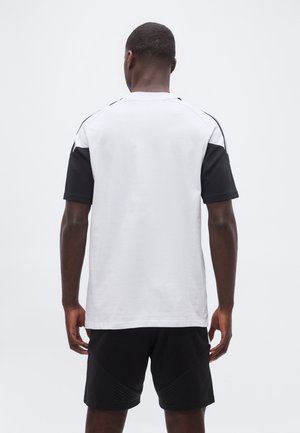 Homme debout de dos, portant un T-shirt blanc à manches courtes noires et un short noir sur un fond uni.