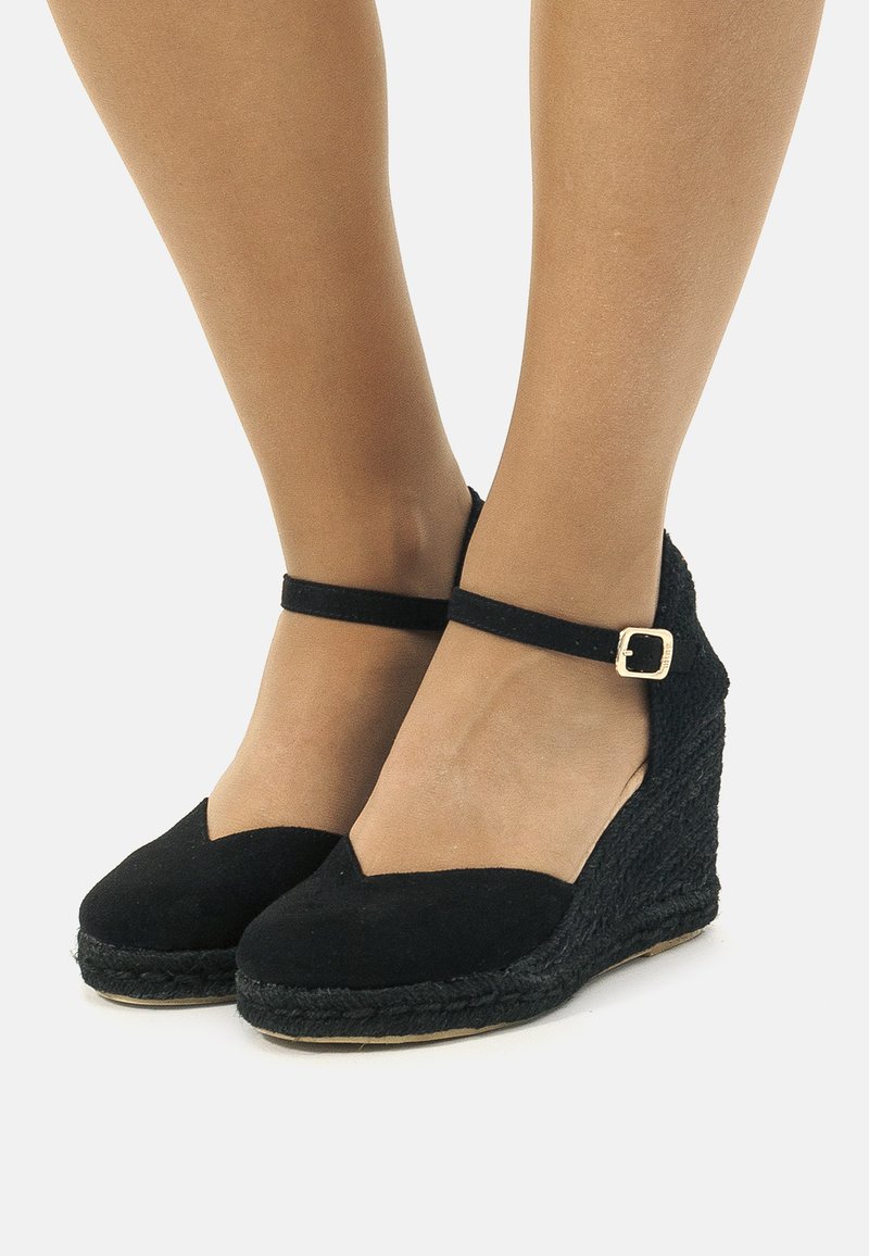 Scarpe con zeppa in suede nero con cinturino alla caviglia, dotate di suola in juta intrecciata e design a forma di V sulla parte anteriore. Il cinturino presenta una piccola fibbia dorata.