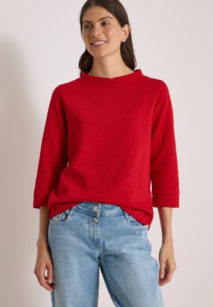 Femme souriante, portant un haut rayé rouge à manches longues et un jean bleu clair, debout devant un fond clair uni.