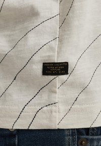 Witte stof met zwarte diagonale stikdetails. Label zegt "FREEDOM UNLIMITED," wat aangeeft dat het is getest op hoge hoogte. Textuur op het oppervlak.