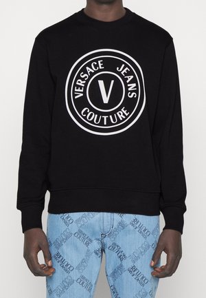 Schwarzer Pullover mit weißem „Versace Jeans Couture“-Kreislogo, getragen mit hellblauen Jeans mit wiederholtem „Versace Jeans Couture“-Print.