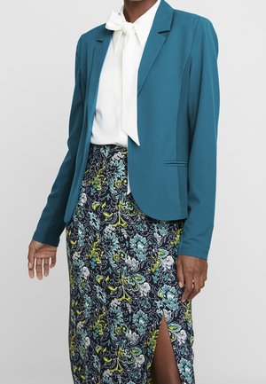 Frau trägt einen türkisfarbenen Blazer, weiße Bluse mit Schleife und einen Rock mit Blumenmuster und vorderem Schlitz.