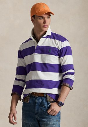 Chemise de rugby à manches longues à rayures blanches et violettes, dotée d'un col et d'un logo vert. Associée à un jean bleu déchiré et une casquette orange.