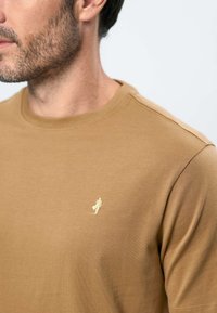 Homme avec une légère barbe portant un t-shirt marron à col rond avec un petit logo blanc brodé sur le côté gauche de la poitrine.