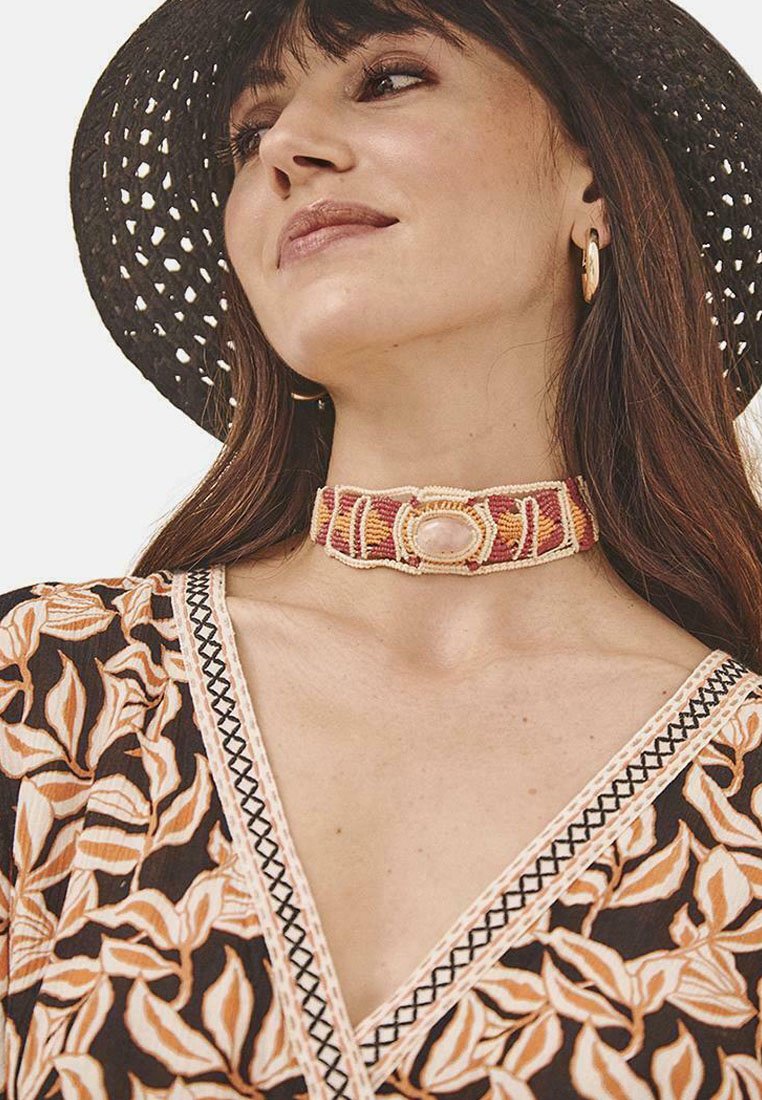 Collar de tela tejido con acentos en rosa y beige, que presenta una piedra ovalada central, combinado con una prenda con patrón floral y un sombrero de ala ancha negro.