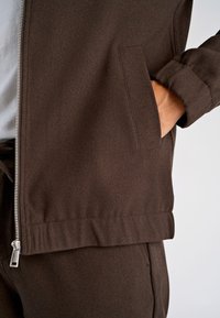 Bruine jas met ritssluiting gemaakt van textuurstof, met een zijzak en een losse pasvorm met een geribde zoom en manchetten.