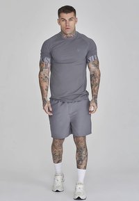 Grå atletisk skjorte og shorts lavet af glat stof. Skjorten har en logo på brystet og mønstrede ærmer. Skoene er hvide med accenter.