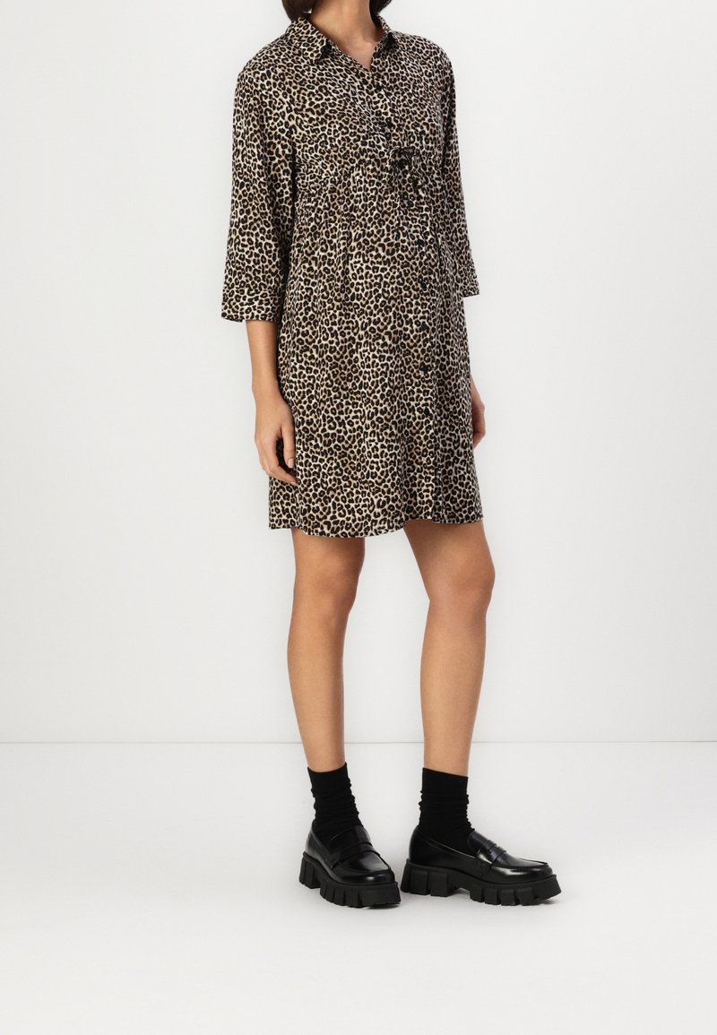 Femme portant une robe mi-longue à imprimé léopard avec des manches trois-quarts, des chaussettes noires et des mocassins noirs épais sur un fond blanc uni.