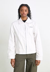 Balta zip-up striukė su vertikaliomis tamsiai mėlynomis juostelėmis, užskaitytu apykaklę ir šoninėmis kišenėmis. Adidas logotipas yra išsiuvinėtas ant krūtinės.