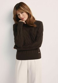 Brązowy sweter dzianinowy z wzorem w warkocze, z prążkowanymi mankietami i dołem, w połączeniu z białymi spodniami. Modelka stoi w zrelaksowanej pozie.