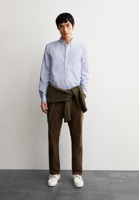 Gestreepte blauwe overhemd, bruine broek, groene trui om de taille geknoopt, en witte schoenen. Glad materiaal met een relaxte pasvorm.