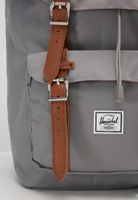 Un sac à dos en tissu gris avec des sangles en cuir marron et des accessoires en métal argenté. Comprend un patch logo Herschel à l'avant.