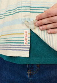 Gestreepte shirt in crème, blauw en geel met een label waarop "REVOLUTION" staat. Gelaagd over een teal T-shirt, met denim zichtbaar eronder.
