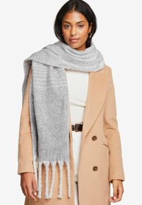 Écharpe en tricot gris avec des bords frangés posée sur les épaules, présentant une texture douce et des motifs subtils, associée à un manteau beige.
