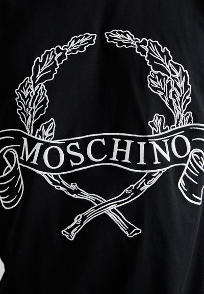 Μαύρος ύφασμα με λευκό κεντητό σχέδιο που περιλαμβάνει δάφνινες κλαδιά και σταυρωτά ραβδιά, μαζί με το κείμενο «Moschino» σε έντονα γράμματα.