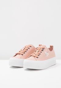 Ljusrosa canvas-sneakers med vit plattformssula, prydda med runda snören och en logodetalj på sidan. Slät textur, avslappnad design.