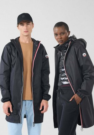 Twee modellen met zwarte hooded jassen met rode en witte ritsen; de een in lichtblauwe jeans en een beige shirt, de ander in zwarte broek en een glanzend vest.