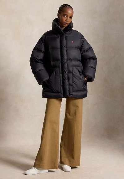 Kvinde iført en sort dunjakke, lysebrune vidde bukser og hvide sko, stående med hænderne i lommerne mod en neutral baggrund.