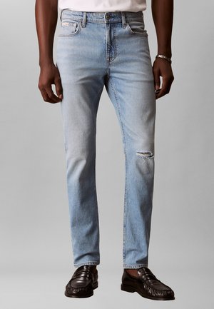 Uomo che indossa jeans slim fit azzurri con una piccola lacerazione sul ginocchio destro e mocassini in pelle nera, in piedi contro uno sfondo semplice.