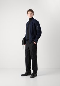 Jūras spēku bumbvedēju jaka ar rāvējslēdzēju, apvienota ar melnu turtleneck, kraukšķīgajām biksēm un melnām kurpēm. Modelis tur nelielu melnu somu.