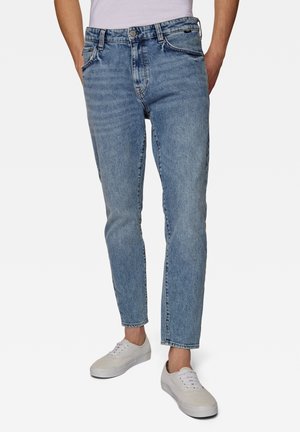 Persoon draagt lichtblauwe denim jeans met een rechte pasvorm, gecombineerd met witte lage sneakers en een ingestopt wit overhemd.
