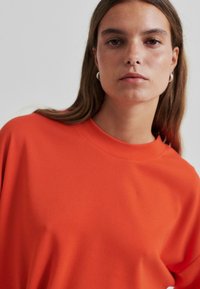 Oranžová oversized mikina s širokým výstřihem, hladkým povrchem a uvolněným střihem. Obsahuje viditelné švy a měkkou tkaninu.