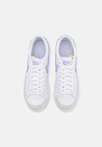White sneakers with purple accents, featuring a smooth leather upper, mesh tongue, and rubber sole. Laces are light pink. Branding is visible.