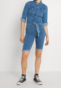 Person som bär en åtsittande denimskjorta med knappar och uppvikta ärmar, tillsammans med matchande denimshorts med en knuten bälte, parat med svarta högklackade sneakers.