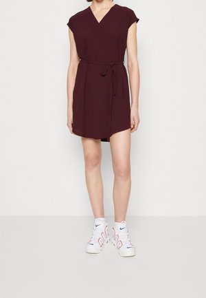 Burgunderfarbenes kurzes Kleid mit V-Ausschnitt, kurzen Ärmeln, einem gebundenen Taillengürtel und einem strukturierten Stoff. Getragen mit weißen Sneakers.