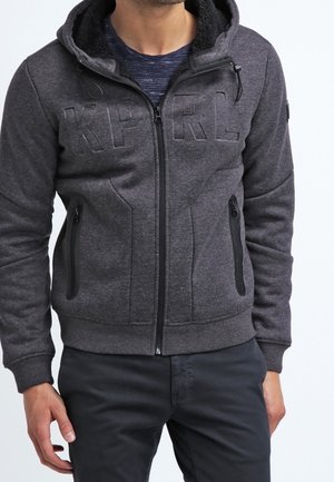 Mann trägt dunkelgrauen Kapuzenpullover mit Reißverschluss und Fronttaschen sowie erhabenen Buchstaben "KP RL" auf der Brust, kombiniert mit schwarzer Hose.