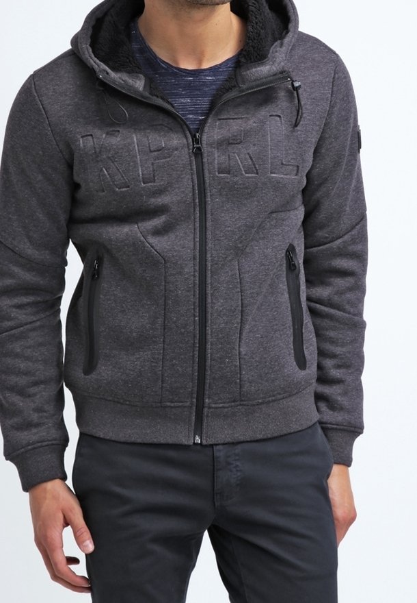 Homme portant un sweat à capuche zippé gris foncé avec poches avant et lettres en relief "KP RL" sur la poitrine, associé à un pantalon noir.