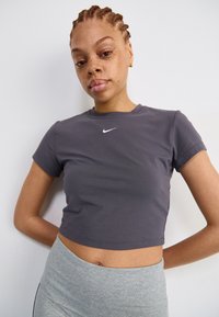 Grijze katoenen crop top met een ronde halslijn en korte mouwen, met een wit Nike-logo op de borst. Huidskleurige leggings zichtbaar bij de taille.
