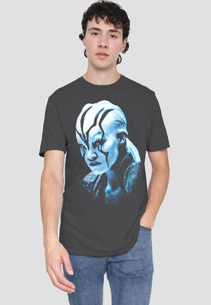 Graues Baumwoll-T-Shirt mit einem blauen Grafikdesign eines Charaktergesichts, das mit tribalähnlichen Mustern verziert ist, vor einem schlichten kreisförmigen Hintergrund.