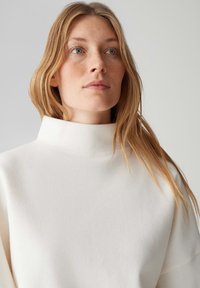 Weißer Pullover mit hohem Kragen, weicher, glatter Textur und minimalistischem Design. Verfügt über lockere Schultern, keine sichtbaren Muster oder Applikationen.