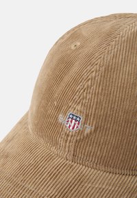 Brun kordflöjtsmössa med en broderad logotyp som har en sköld med röda och vita ränder samt blå stjärnor. Strukturerad och texturerad design.