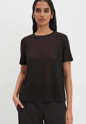 Donna che indossa un top nero semplice a maniche corte con scollo rotondo, la mano sinistra nella tasca dei pantaloni neri, sfondo neutro.