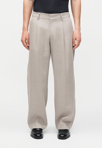 Homme portant un pantalon beige clair à larges jambes à rayures fines avec des plis, associé à des chaussures noires et un haut noir, debout devant un fond uni.