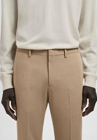 Pantalones beige a medida de tela ligera, con frente plano, trabillas para cinturón y un acabado liso y limpio.