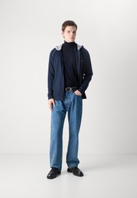 Felpa con zip navy e cappuccio grigio, dolcevita nera, jeans in denim azzurro chiaro e scarpe nere, con una vestibilità ampia e uno stile casual.