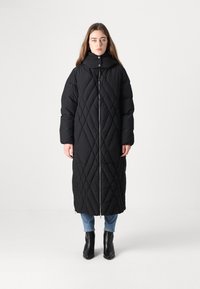 Crni preklopni dugi kaput s visokim ovratnikom i prednjim patentnim zatvaračem. Sadrži dijagonalne uzorke i opuštenu siluetu, uparen s plavim trapericama i crnim čizmama.