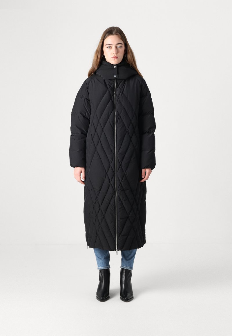 Crni preklopni dugi kaput s visokim ovratnikom i prednjim patentnim zatvaračem. Sadrži dijagonalne uzorke i opuštenu siluetu, uparen s plavim trapericama i crnim čizmama.