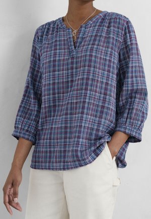Personne portant une chemise à carreaux bleue et rouge à manches longues avec les poignets retroussés et un pantalon blanc, une main dans la poche.