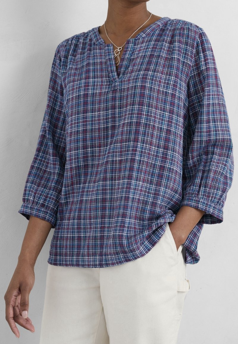 Personne portant une chemise à carreaux bleue et rouge à manches longues avec les poignets retroussés et un pantalon blanc, une main dans la poche.