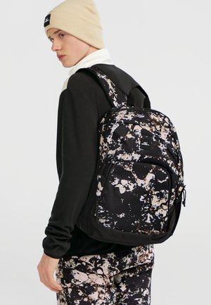Mochila con base negra, que presenta un patrón salpicado en beige y blanco. Incluye correas acolchadas y un bolsillo delantero con cremallera.
